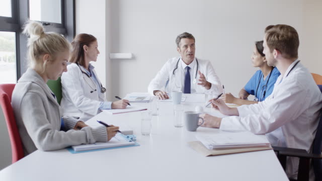 Dolly shot of mature doctor discussing with colleagues in board room. Male and female healthcare workers are sitting at conference table. They are working in hospital.